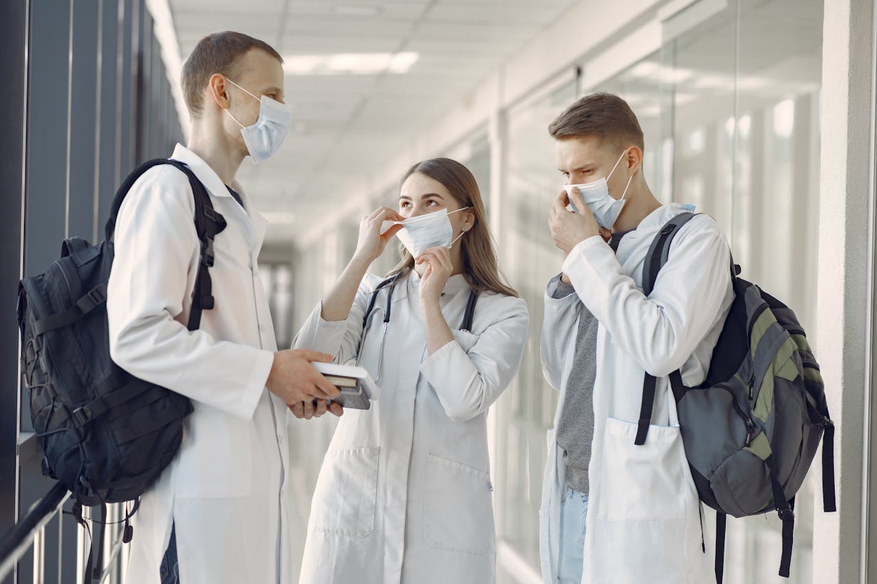 Three young medical professionals discuss in a hospital corridor wearing protective masks.